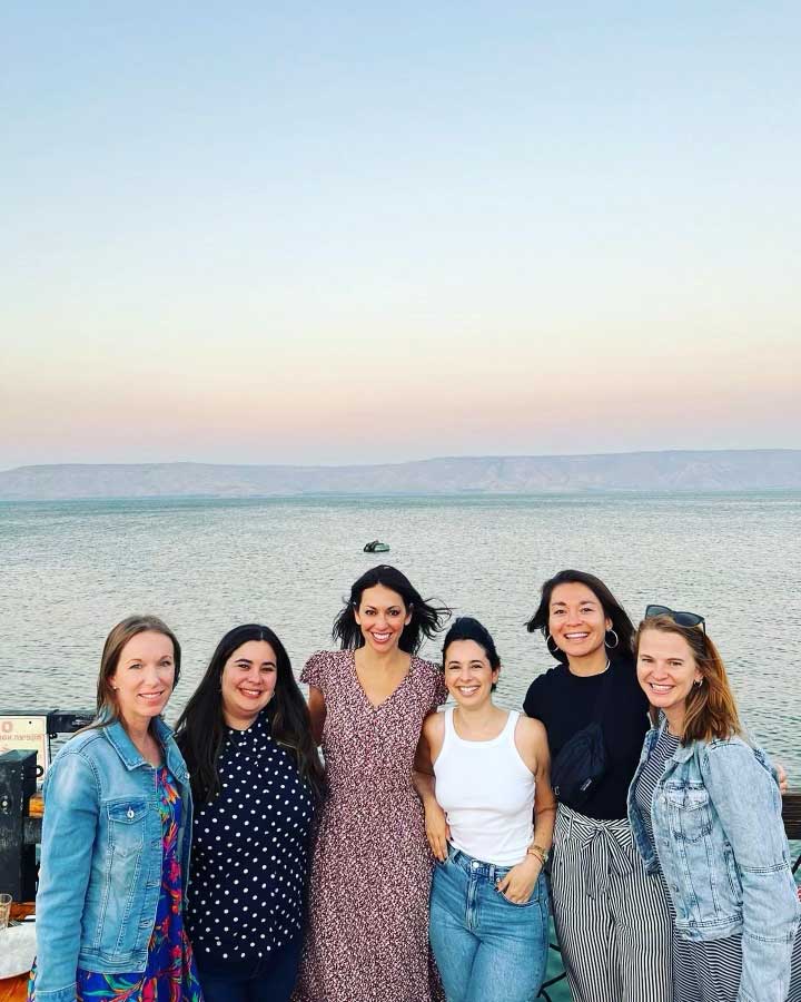 group of six people posing together by a body of water during sunset