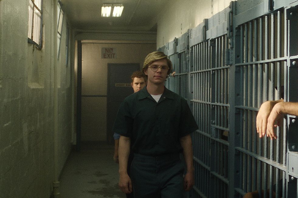 evan peters portrays jeffrey dahmer walking through a jail while wearing a dark teal inmate uniform and aviator glasses, behind him is another actor, and a third actor is shown behind bars on the right
