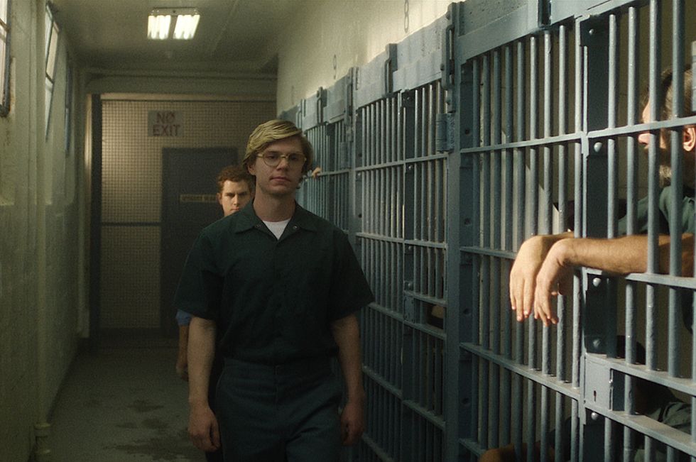evan peters portrays jeffrey dahmer walking through a jail while wearing a dark teal inmate uniform and aviator glasses, behind him is another actor, and a third actor is shown behind bars on the right