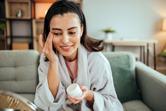 mujer en albornoz poniendose crema en la cara ante un espejo de mano