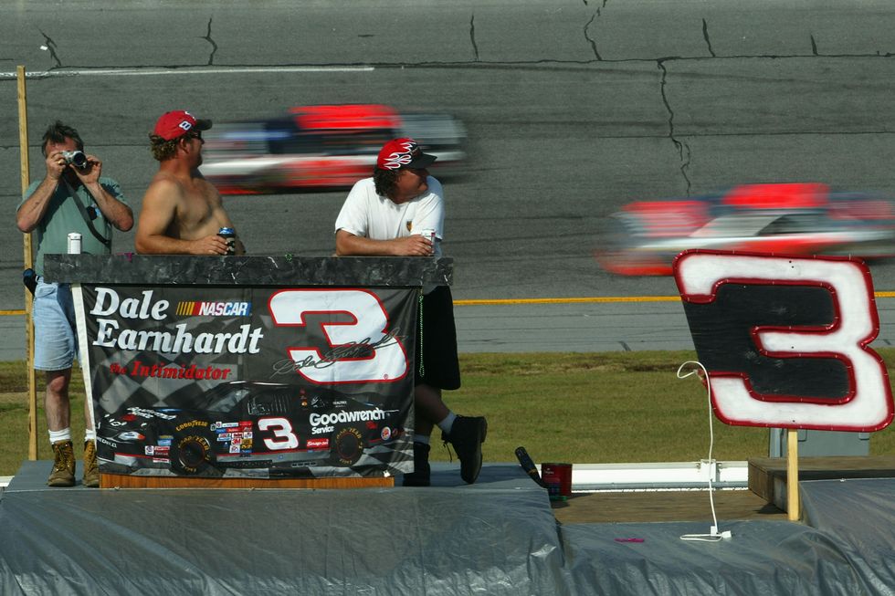 people standing behind a large flag as race cars drive by in the background