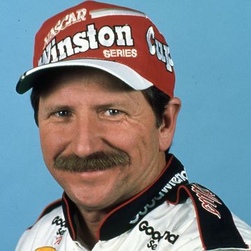 a man in a racing jacket and red ball cap smiles at the camera
