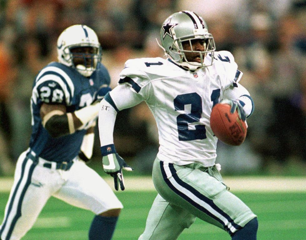 deion sanders runs while holding a football in his left hand, behind him is an opponent running after him, both men wear football uniforms and helmets
