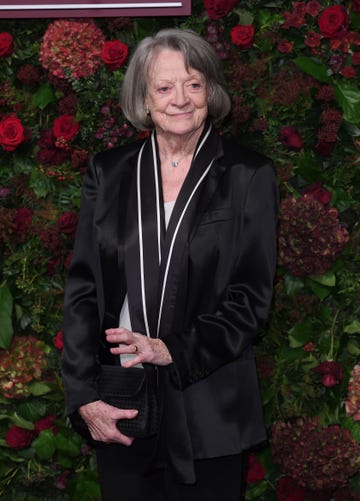 maggie smith smiles while standing in front of a wall of flowers and greenery, she looks too the right and wears a black jacket with white trim