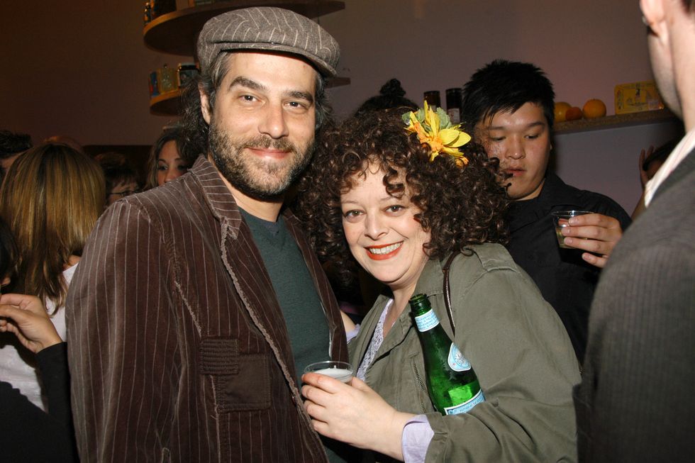danny keough and angela mccluskey embrace and smile for a photo, they stand in a crowded room, he wears a brown blazer and a matching hat, she has a yellow flower in her hair