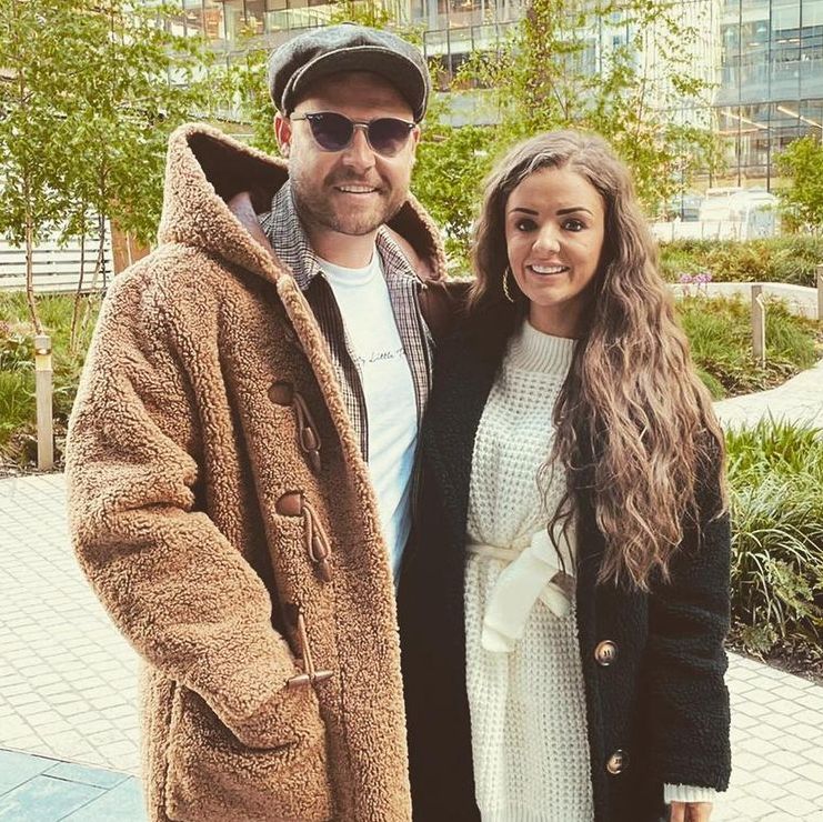 danny miller with fiancee steph jones posing in front of trees and glass building danny wears check trousers, a long furry coat, flat cap and sunglasses, while steph wears a white knitted dress and long black furry coat