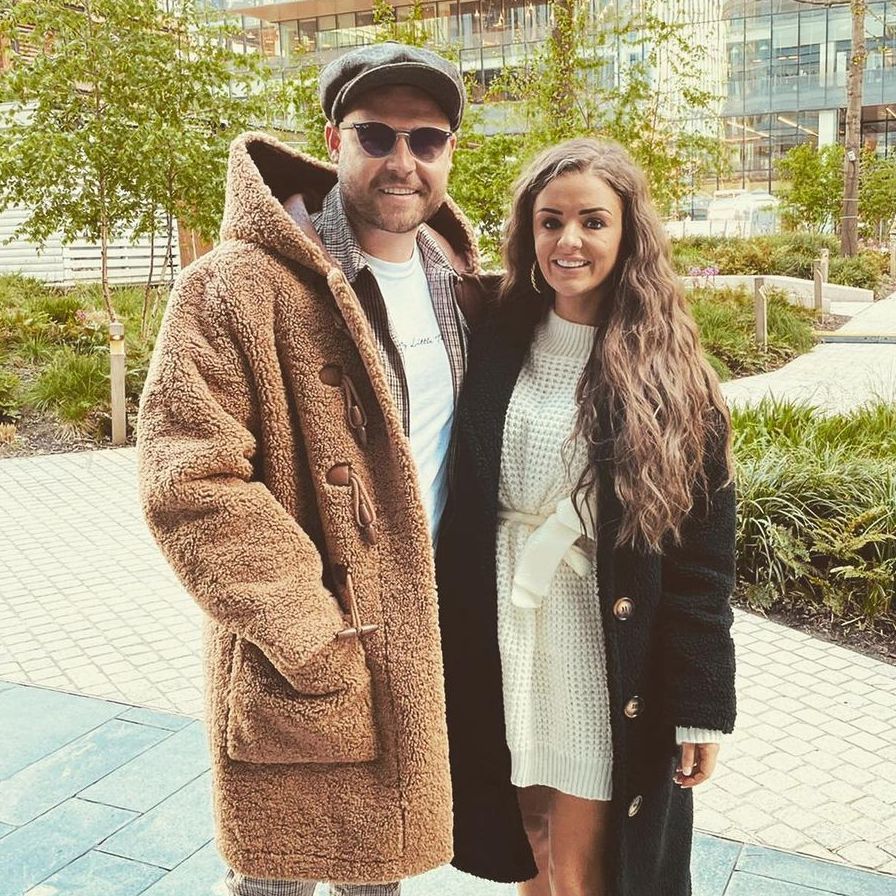 danny miller with fiancee steph jones posing in front of trees and glass building danny wears check trousers, a long furry coat, flat cap and sunglasses, while steph wears a white knitted dress and long black furry coat