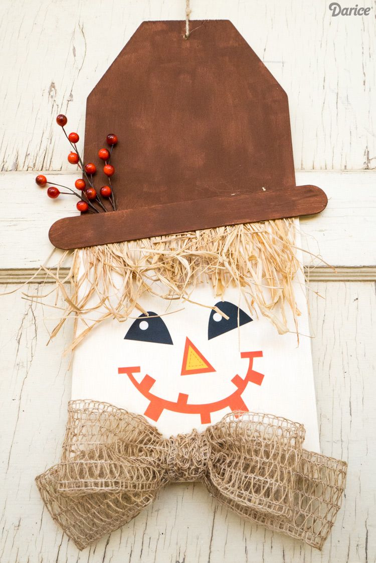 wooden scarecrow door hanger with berry spray on hat, hair made of raffia, and bow made of burlap like ribbon