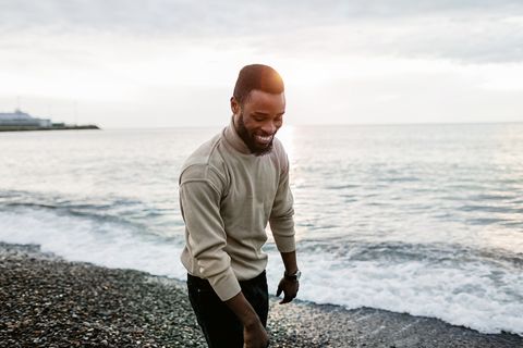 young man with black hair, beard and mustache walks along the beach along the sea and smiles and looks down