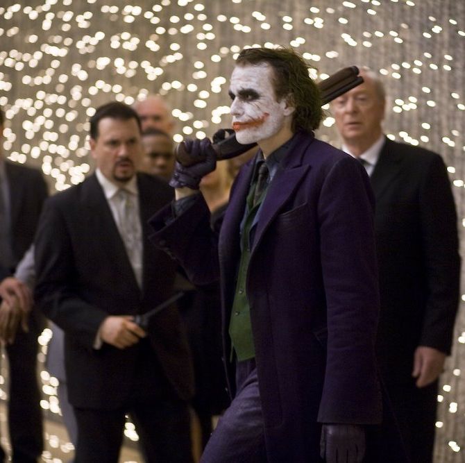 the dark knight   medium shot of heath ledger as joker at party holding gun with crowd in background including michael caine as alfred