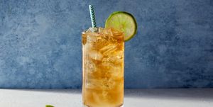 glass of a dark and stormy cocktail with a paper straw and a lime sitting on a stone surface with a marbled blue background