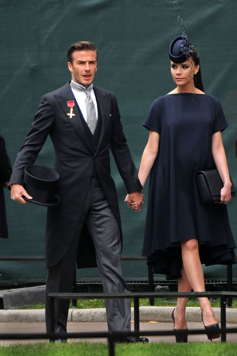 david beckham and victoria beckham walk hand in had on a sidewalk, he wears a dark three piece suit with a long jacket and holds a top hat, she wears a navy dress and matching hat