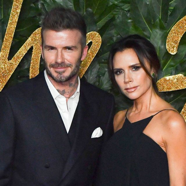 david and victoria beckham pose for a photo while standing in front of a green leaf background, he wears a black suit and she wears a black dress