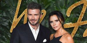 david and victoria beckham pose for a photo while standing in front of a green leaf background, he wears a black suit and she wears a black dress
