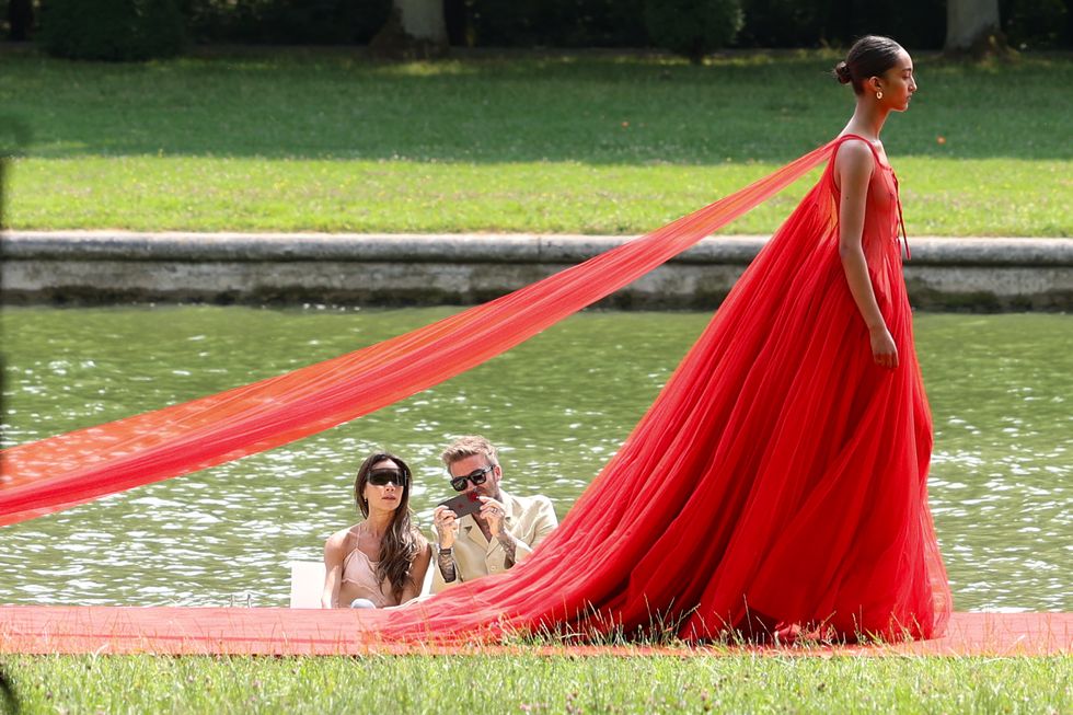 victoria and david beckham watch a fashion show while sitting in a boat, a model in an avant garde red tulle dress walks in front of them