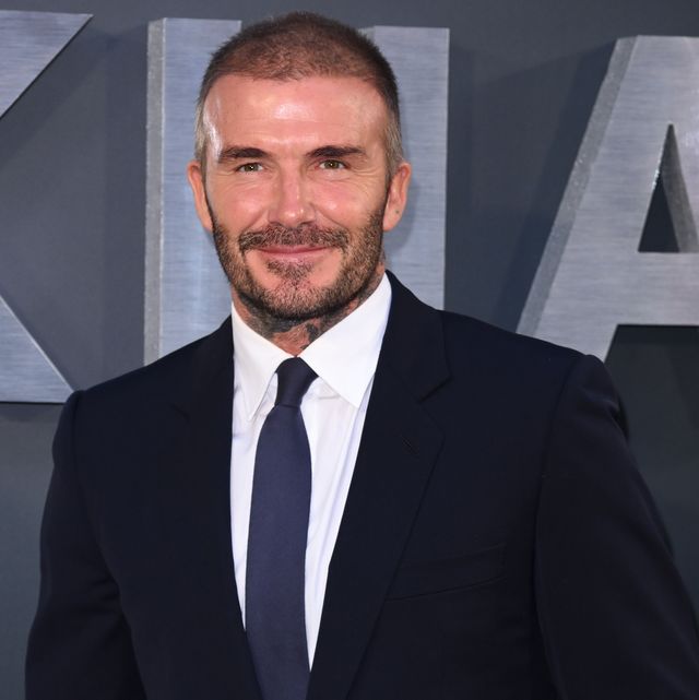 david beckham smiles at the camera, he wears a dark suit jacket, white collared shirt and navy blue tie