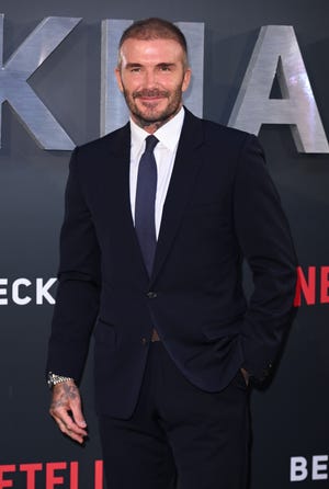 david beckham smiles at the camera, he wears a dark suit jacket, white collared shirt and navy blue tie