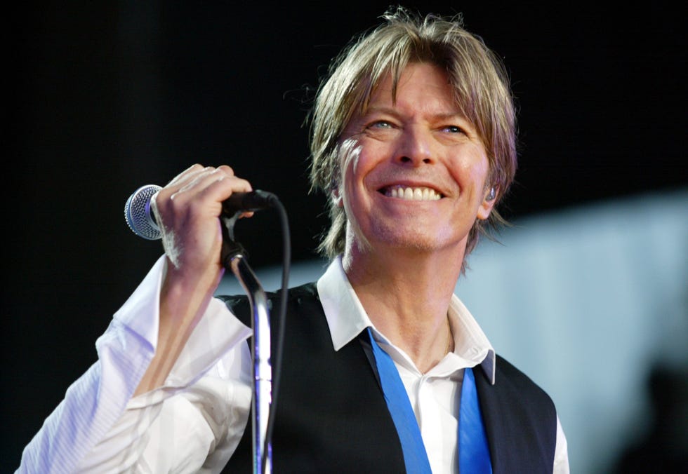 david bowie smiles and looks past the camera as he grips a microphone on a stand in one hand