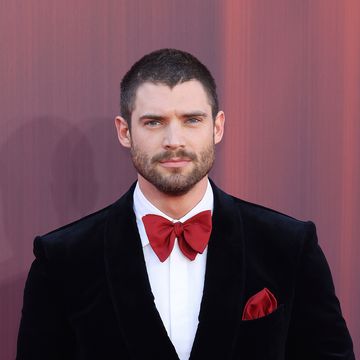a man stares at the camera while standing in front of a rust red colored background, he wears a black suit coat white collared shirt red bowtie and red pocket square