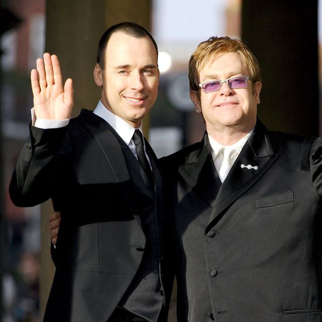 david furnish and elton john both wave and smile, they are both wearing black suits with white collared shirts and ties, elton wears silver framed sunglasses with purple tinted lenses
