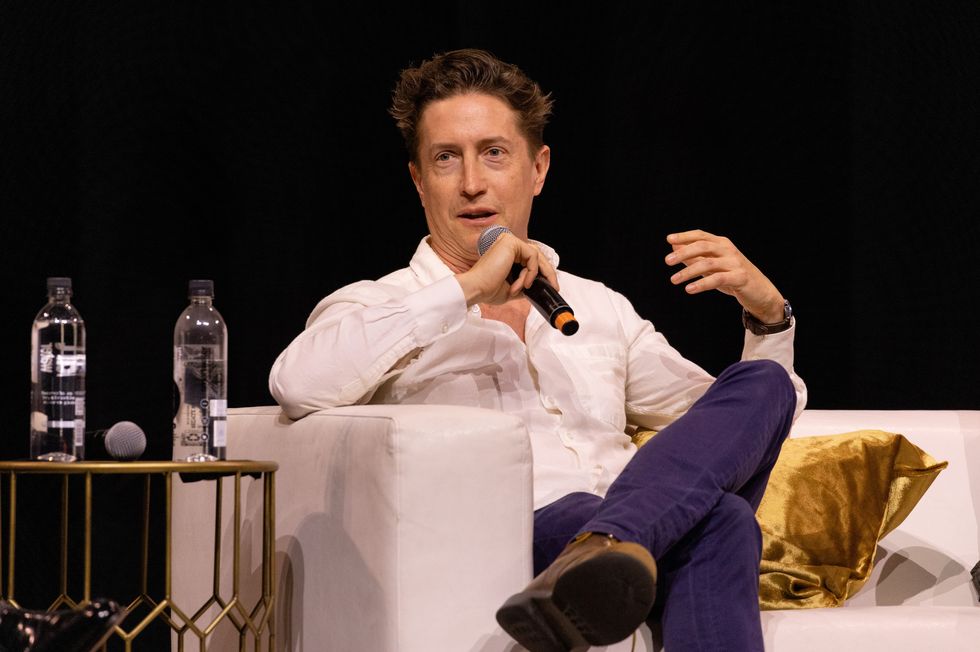 David Gordon Green, un hombre vestido con una camisa blanca con botones y pantalones morados, sosteniendo un micrófono mientras habla en el escenario.