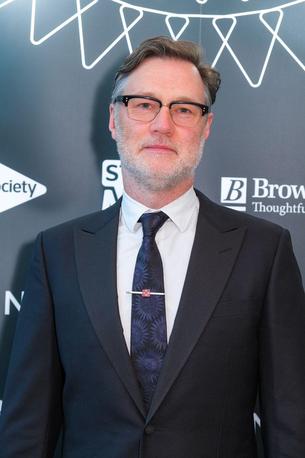 David Morrissey, un hombre mayor mira a la cámara con una expresión facial neutra, lleva gafas con el pelo canoso peinado hacia atrás, chaqueta de traje negra, camisa blanca y corbata azul.