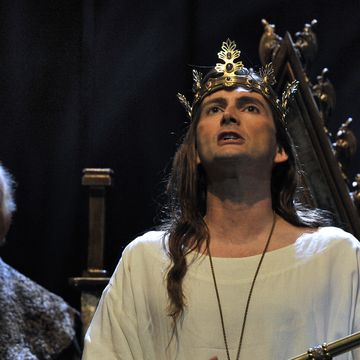 david tennant, as richard ii, stands in front of a throne wearing a crown and robe and holding a sceptre as he looks upwards