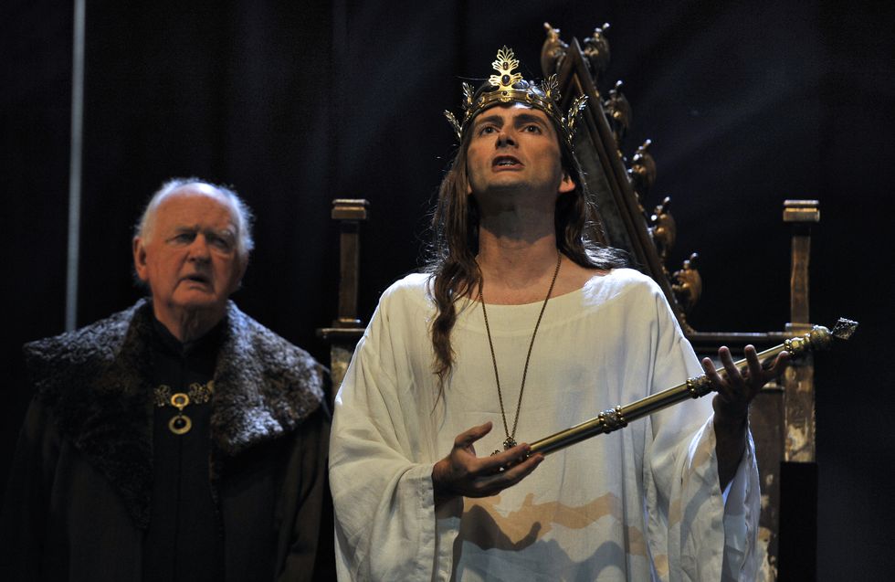 David Tennant steht als Richard II. vor einem Thron, trägt eine Krone und ein Gewand, hält ein Zepter und blickt nach oben