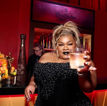 davine joy randolph holds a glass to the camera as she leans against a bar, an oscar trophy, flowers and a glittery bottle sit on the bar next to her, she wears a black beaded strapless dress