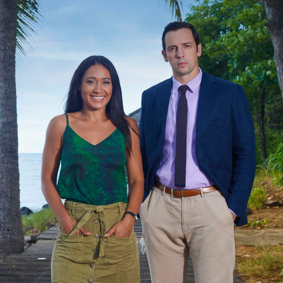 detectives florence cassell and neville parker stand next to each other on a beach in death in paradise