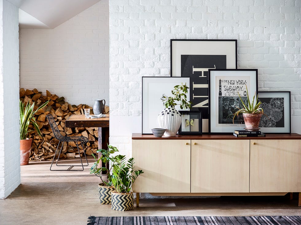 arrangement of prints and artwork, plants on retro style wood sideboard, black and grey stripe pattern rug, houseplants, painted brick wall, concrete floor, view through open doorway to logs stored in room beyond with wooden table and metal chair, neutral, natural materials, textures,