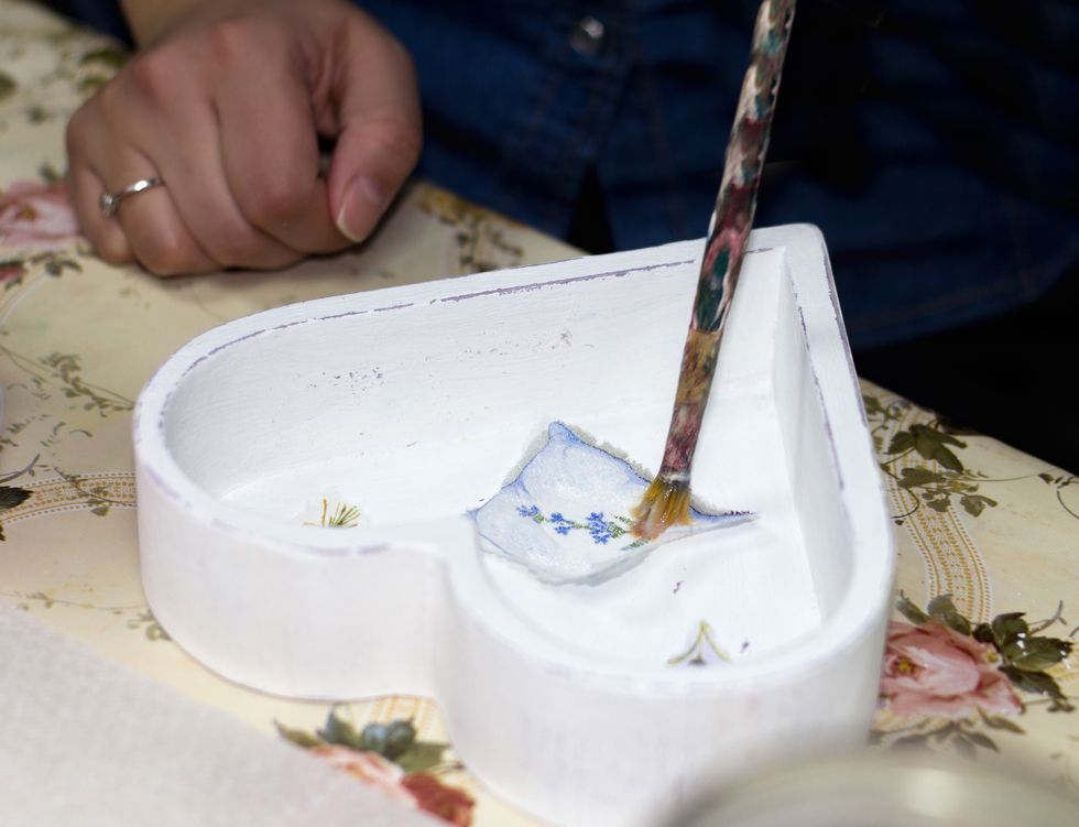an artisan's hand painting a cut out image with glue on inside of a white, heart shaped box