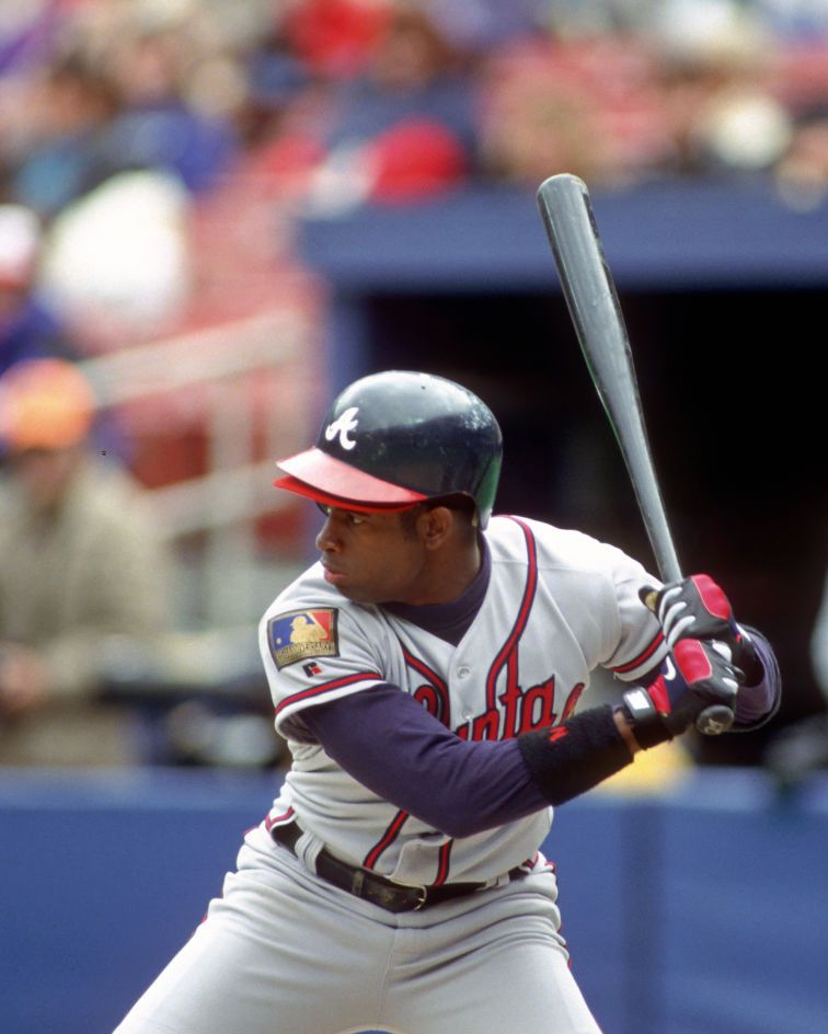 deion sanders holds a bat as he looks forward preparing to hit, he wears a light gray atlanta braves uniform with a blue hitting cap and red and black gloves