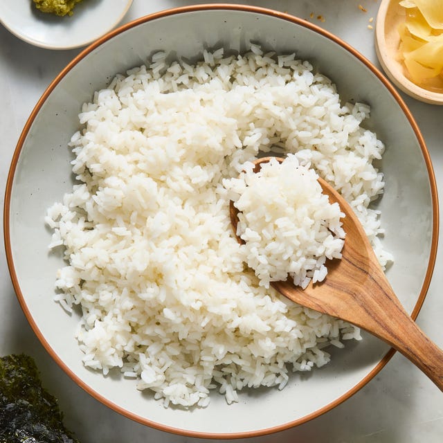 sushi rice in a bowl with a wooden spoon and bowls of carrots, cucumber, pickled ginger, and wasabi