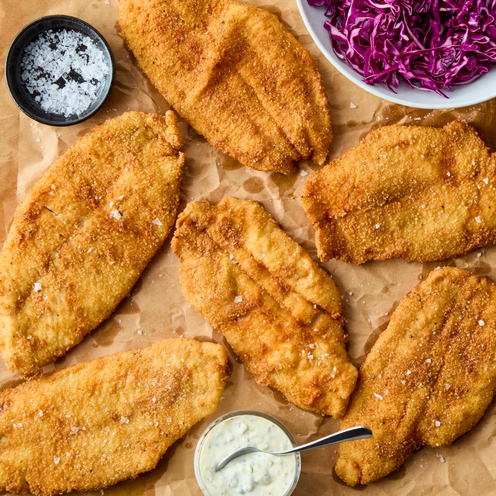 matzo fried fish on brown paper with a bowl of red cabbage, a bowl of flaky salt, and a bowl of tarter sauce