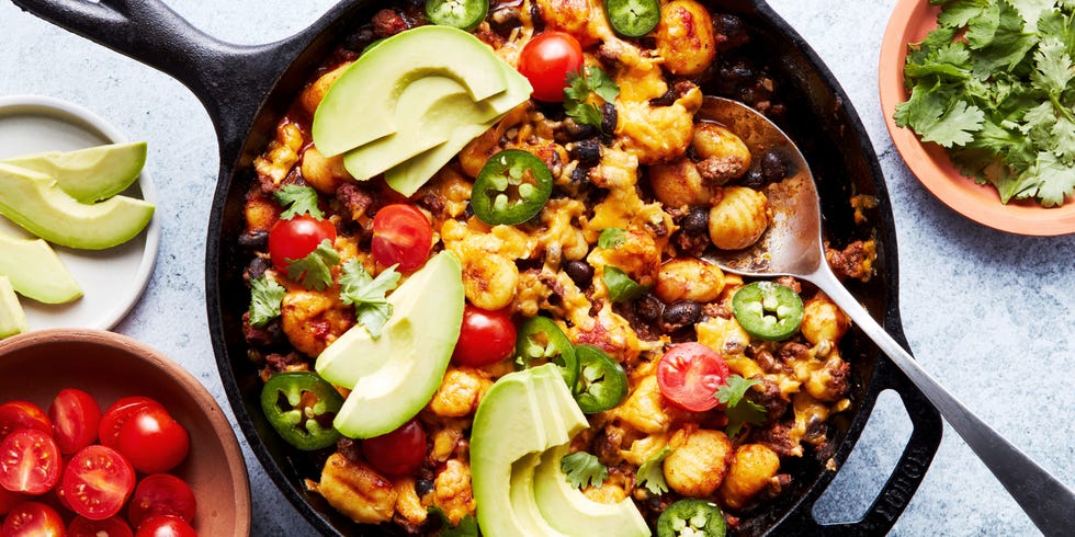 a pan with gnocchi covered in enchilada sauce, cheese, avocado, cilantro, and cherry tomatoes