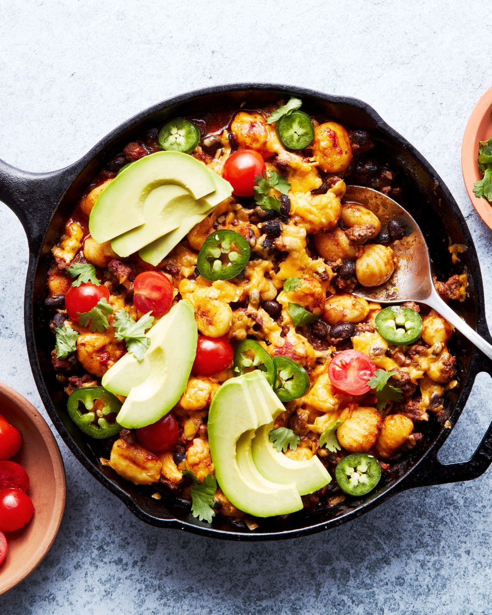 a skillet with gnocchi, ground beef, enchilada sauce, cheese, avocado, jalapeno, cilantro, and cherry tomatoes