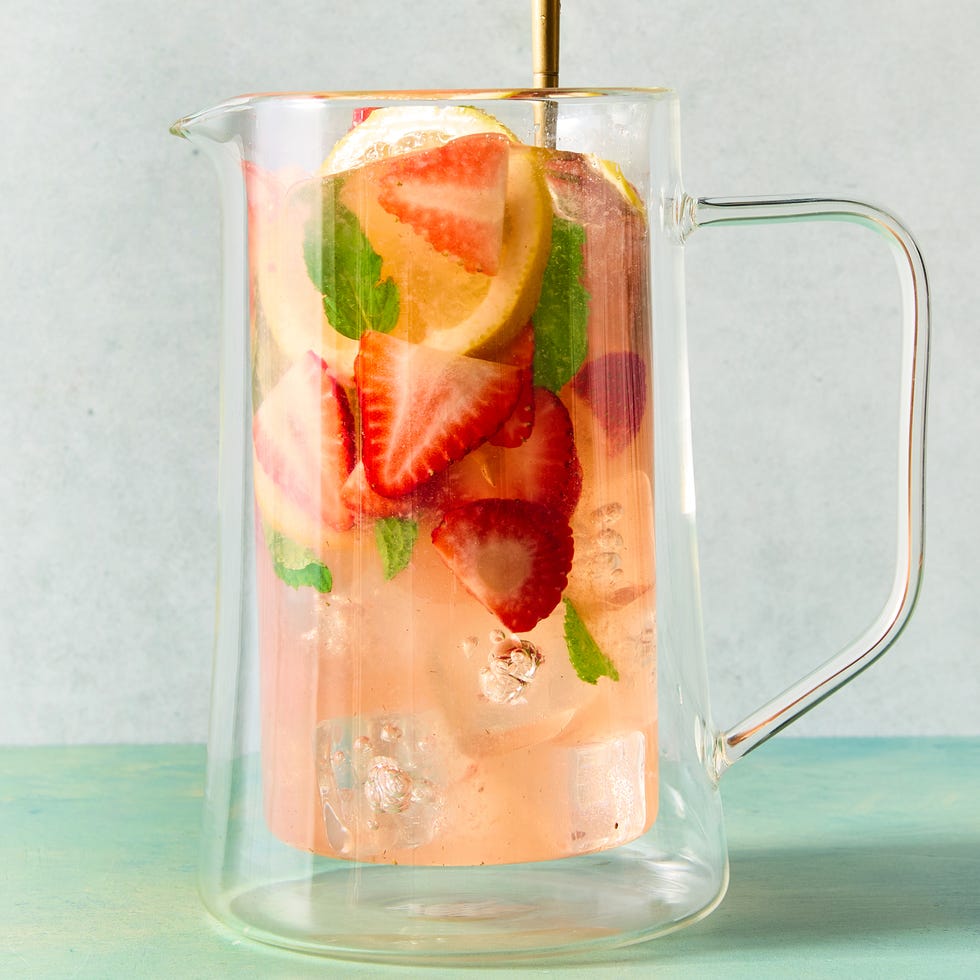 a pitcher and glasses of sangria made with white wine and strawberry syrup with ice, pieces of mint, sliced strawberry, and sliced lemons