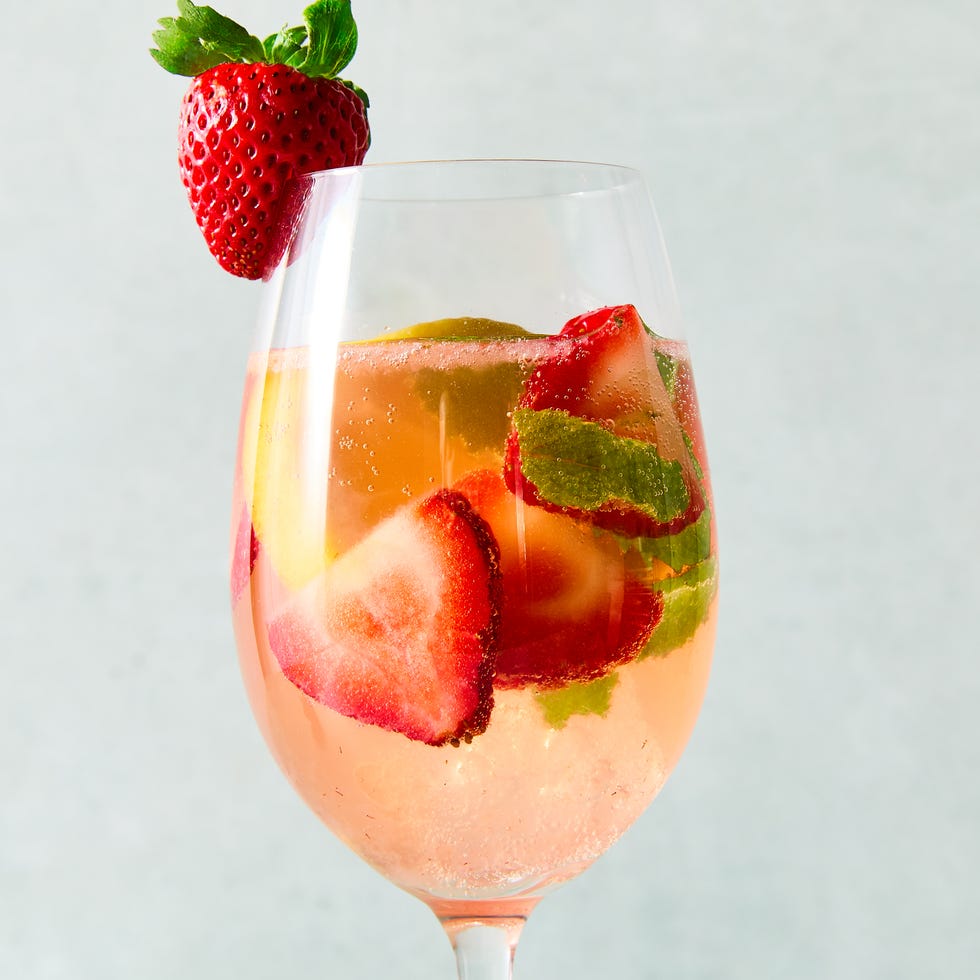 a pitcher and glasses of sangria made with white wine and strawberry syrup with ice, pieces of mint, sliced strawberry, and sliced lemons