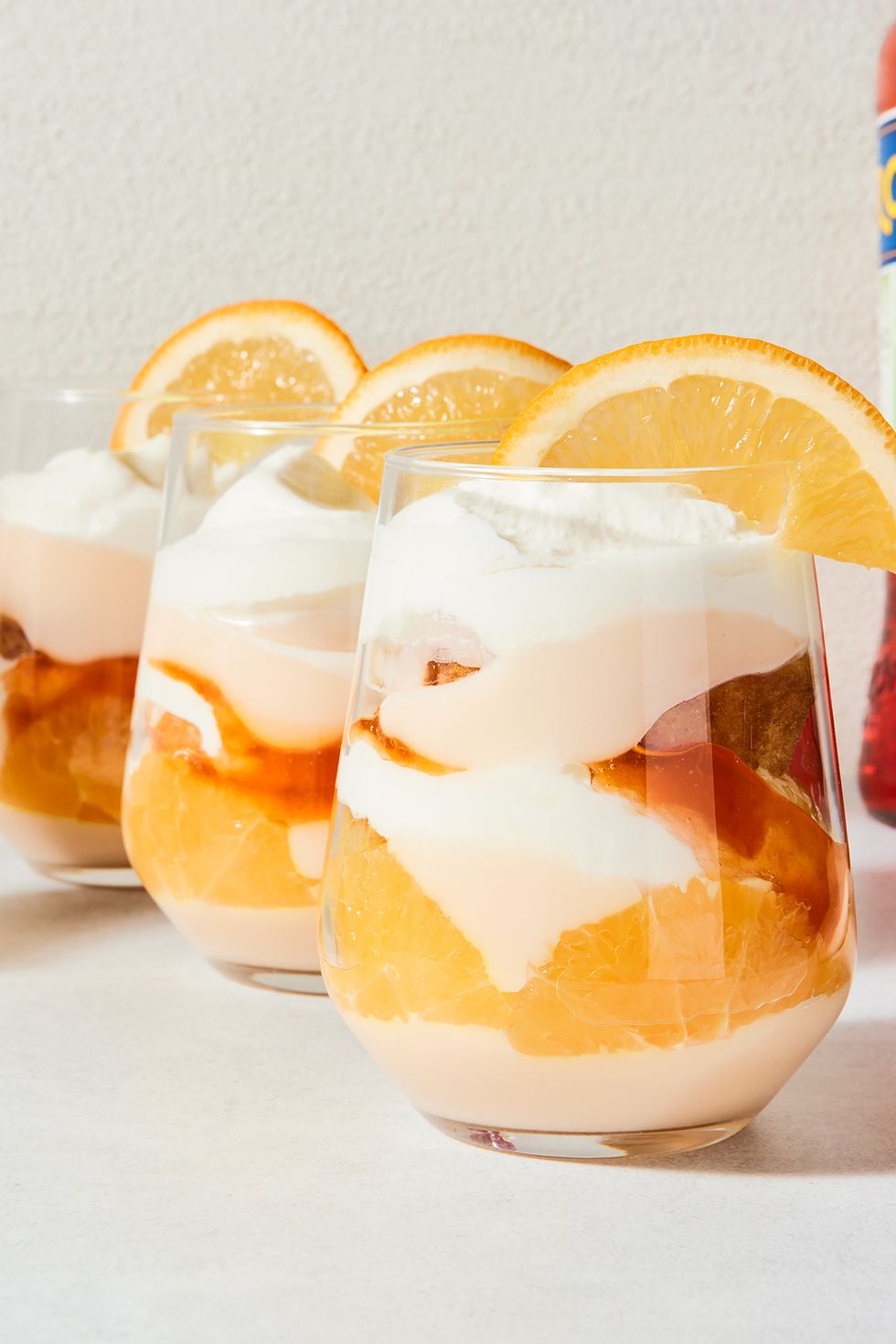 a stemless wine glass filled with vanilla cake, whipped cream, aperol spiked jam, and pudding with an orange slice on the rim