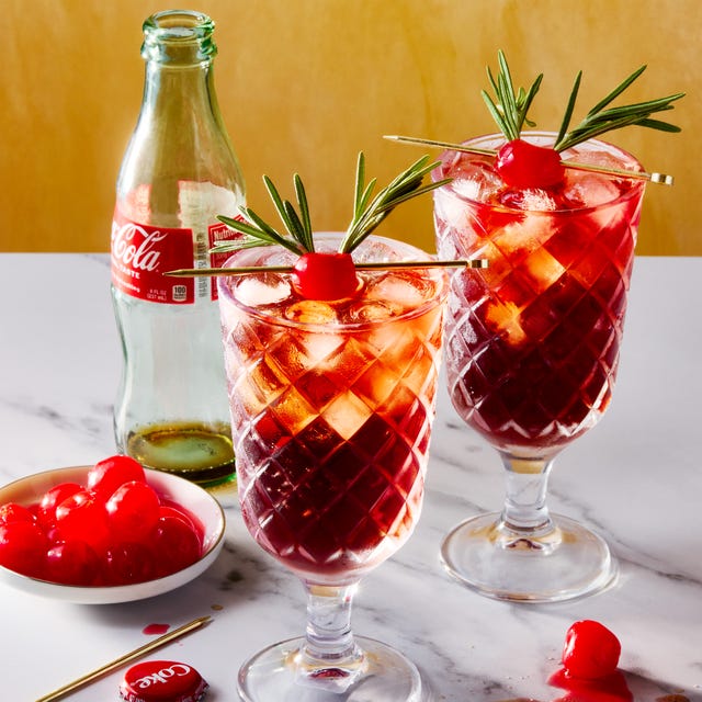 two glasses filled with ice, rum, cherry juice, and cola, garnished with a maraschino cherry decorated with two sprigs of rosemary