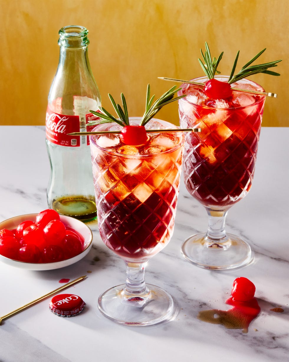 two glasses filled with ice, rum, cherry juice, and cola, garnished with a maraschino cherry decorated with two sprigs of rosemary