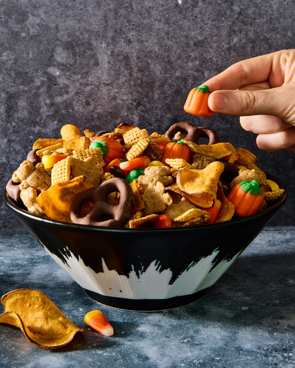 a bowl of halloween themed chex mix with chex cereal, chocolate pretzels, reese's pieces, candy corn, and sweet potato chips