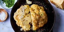 whole roasted cauliflower in a cast iron skillet beside bowls of chopped parsley and salt and a block of cheese