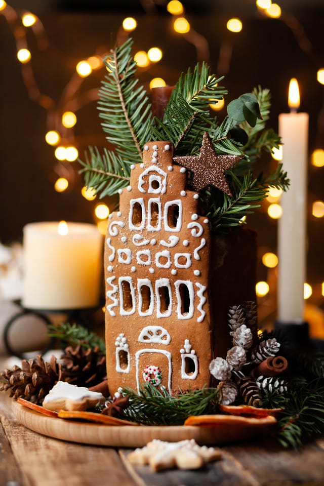 delicious homemade baked gingerbread house with white icing rustic style, white wooden background, candles and christmas lights as decor cozy home atmosphere, festive mood, holiday inspiration