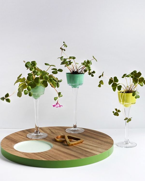 There are three planters made of tall glass candle holders, each with a cup painted a different shade of green and a clover inside.