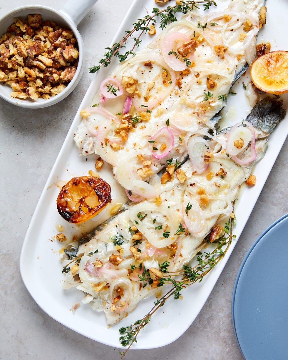 whole roasted trout filets on a white platter garnished with fresh thyme, roasted crushed walnuts, and roasted shallots charred lemons on either side of fish empty blue plate in bottom right corner