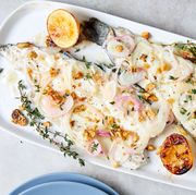 whole roasted trout filets on a white platter garnished with fresh thyme, roasted crushed walnuts, and roasted shallots charred lemons on either side of fish empty blue plate in bottom right corner