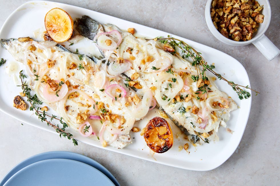 whole roasted trout filets on a white platter garnished with fresh thyme, roasted crushed walnuts, and roasted shallots charred lemons on either side of fish empty blue plate in bottom right corner