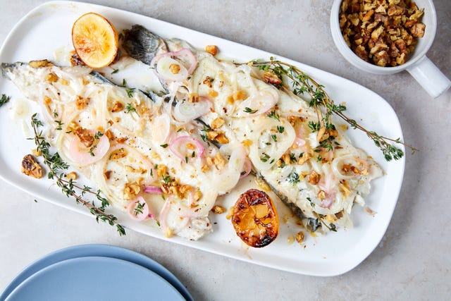 whole roasted trout filets on a white platter garnished with fresh thyme, roasted crushed walnuts, and roasted shallots charred lemons on either side of fish empty blue plate in bottom right corner